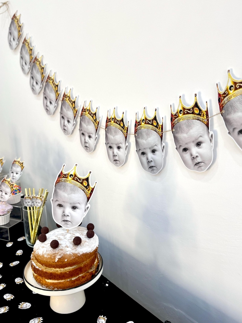 May include: A festive party scene featuring a cake and garland. The cake is topped with a baby's face wearing a gold crown. A garland of baby faces with gold crowns hangs above. The cake is on a white cake stand, and the table is covered with a black tablecloth.