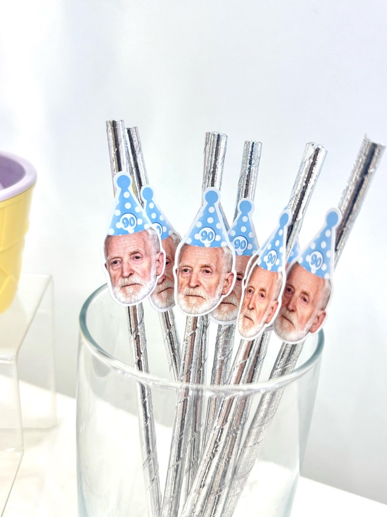 May include: A clear glass filled with silver-colored straws, each topped with a sticker of a man's face wearing a blue party hat with the number 90. The straws are for a birthday celebration.