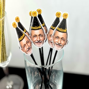 K&ouml;nnte beinhalten: Ein klares Glas gef&uuml;llt mit schwarzen R&uuml;hrst&auml;bchen, die mit Bildern eines l&auml;chelnden Mannes mit einer Partyhut mit der Aufschrift "retired" versehen sind. Die H&uuml;te sind schwarz mit goldenen Verzierungen und einem goldenen Bommel. Die St&auml;bchen befinden sich in einem klaren Glas.