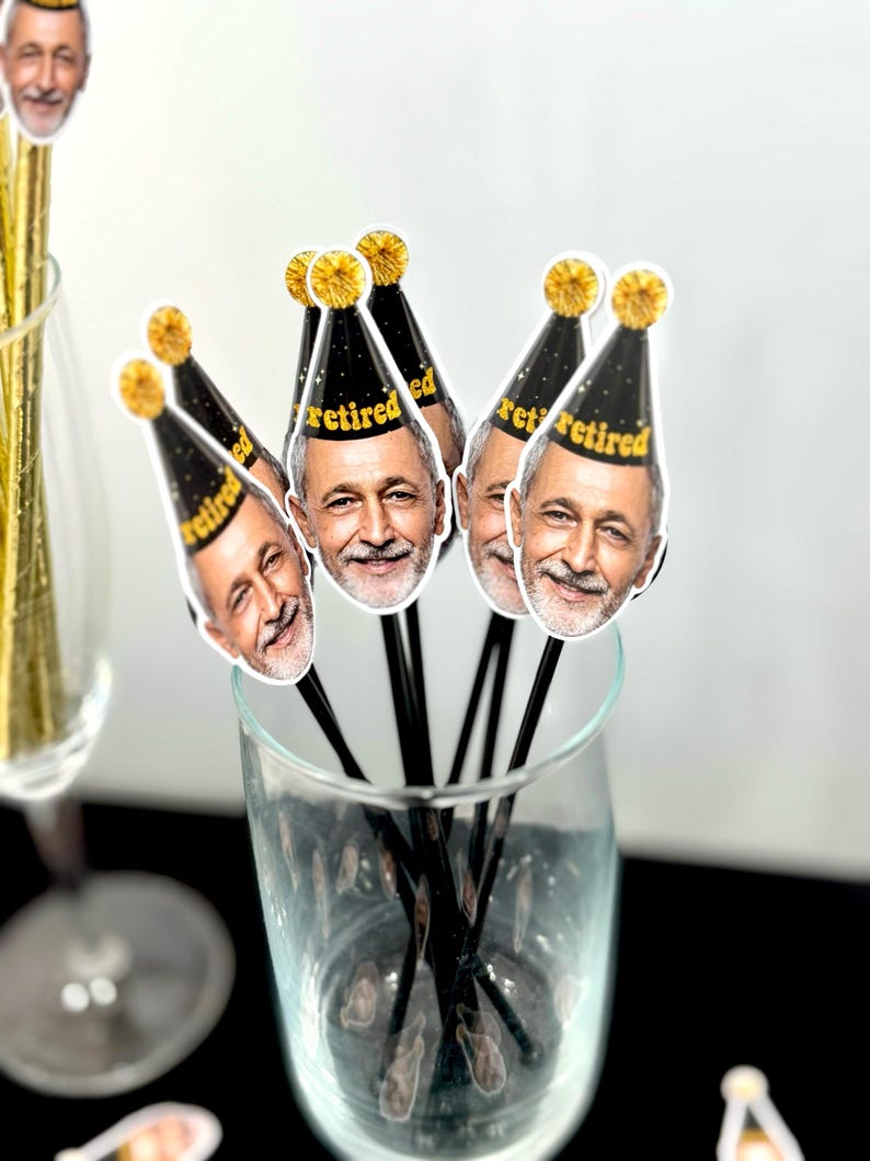 K&ouml;nnte beinhalten: Ein klares Glas gef&uuml;llt mit schwarzen R&uuml;hrst&auml;bchen, die mit Bildern eines l&auml;chelnden Mannes mit einer Partyhut mit der Aufschrift "retired" versehen sind. Die H&uuml;te sind schwarz mit goldenen Verzierungen und einem goldenen Bommel. Die St&auml;bchen befinden sich in einem klaren Glas.