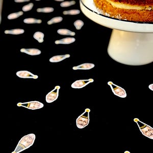 May include: A multi-layered cake on a white cake stand against a black backdrop, decorated with champagne bottle and face stickers. The cake has a white frosting layer and a white and black rimmed plate.