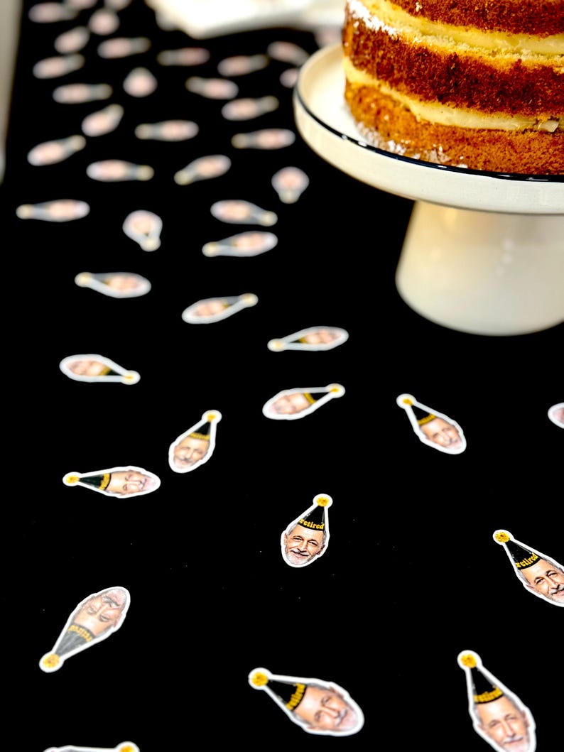 May include: A multi-layered cake on a white cake stand against a black backdrop, decorated with champagne bottle and face stickers. The cake has a white frosting layer and a white and black rimmed plate.