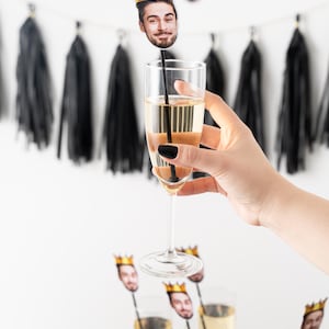 K&ouml;nnte beinhalten: Champagnergl&auml;ser mit Gesichts- und Kronen-Getr&auml;nker&uuml;hrern. Eine Hand h&auml;lt eine Fl&ouml;te mit einem R&uuml;hrer. Ein Banner mit Gesichtern und Kronen h&auml;ngt im Hintergrund. Schwarze Quastengirlande und eine schwarz-wei&szlig;e Tischdecke sind ebenfalls sichtbar.