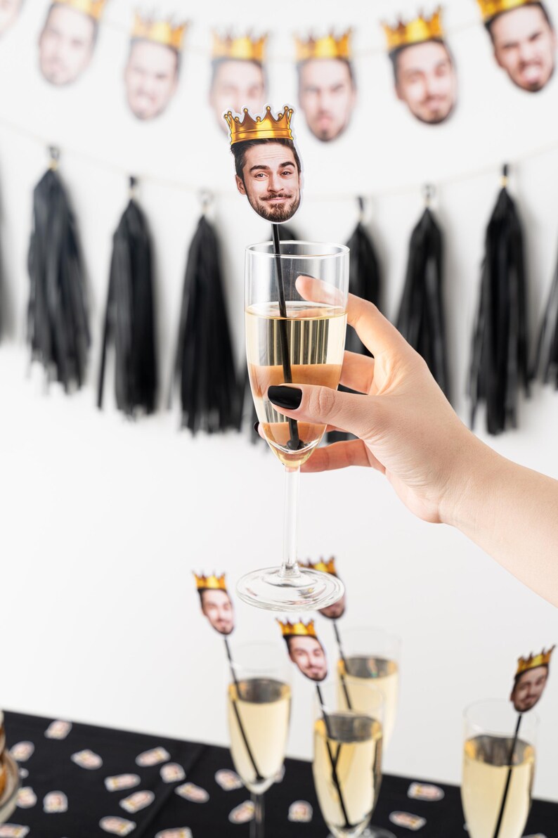 K&ouml;nnte beinhalten: Champagnergl&auml;ser mit Gesichts- und Kronen-Getr&auml;nker&uuml;hrern. Eine Hand h&auml;lt eine Fl&ouml;te mit einem R&uuml;hrer. Ein Banner mit Gesichtern und Kronen h&auml;ngt im Hintergrund. Schwarze Quastengirlande und eine schwarz-wei&szlig;e Tischdecke sind ebenfalls sichtbar.