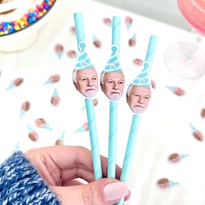 May include: Three light blue paper straws with a printed image of a man wearing a party hat with the number 60. The straws are held in a hand, with a cake and drink in the background. Party supplies.