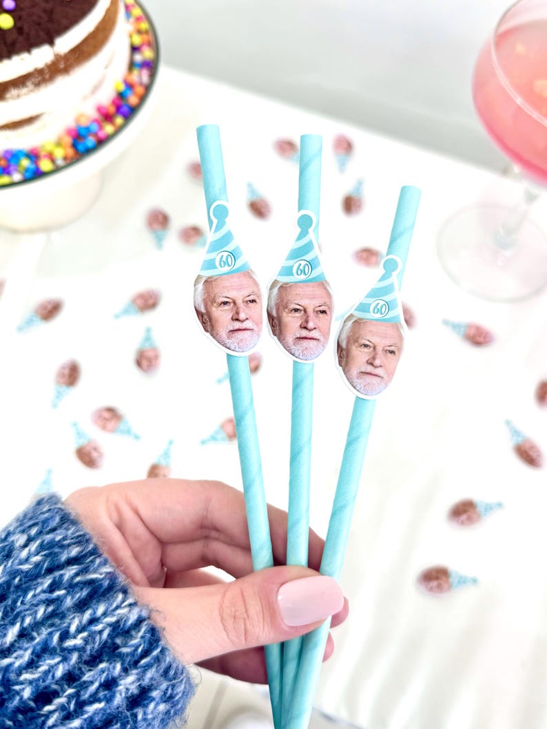 May include: Three light blue paper straws with a printed image of a man wearing a party hat with the number 60. The straws are held in a hand, with a cake and drink in the background. Party supplies.