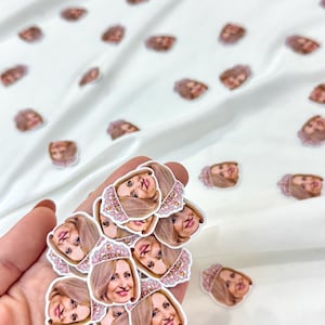 May include: A collection of stickers featuring a smiling woman with blonde hair and a pink tiara. The stickers are scattered on a white surface, with some held in a hand. The stickers have a detailed, illustrated design.