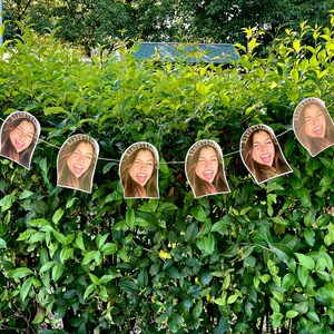 May include: A string of paper cutouts featuring a smiling person with a headband that says "BIRTHDAY". The cutouts are strung together on a white string and are displayed against a backdrop of green foliage.