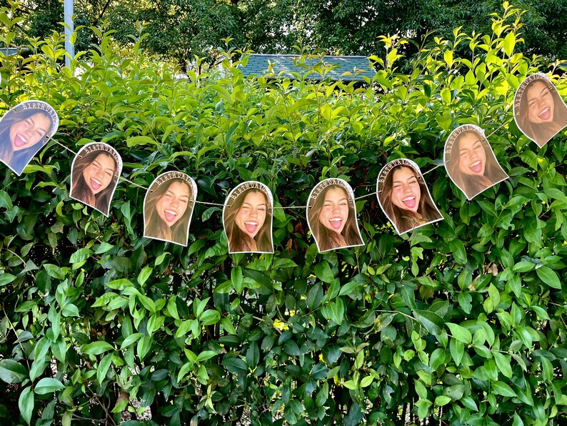 May include: A string of paper cutouts featuring a smiling person with a headband that says "BIRTHDAY". The cutouts are strung together on a white string and are displayed against a backdrop of green foliage.