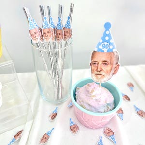 May include: Party decorations featuring a man's face wearing a blue party hat with the number 90. The decorations include drink stirrers in a glass, a cupcake with a topper, and scattered confetti.