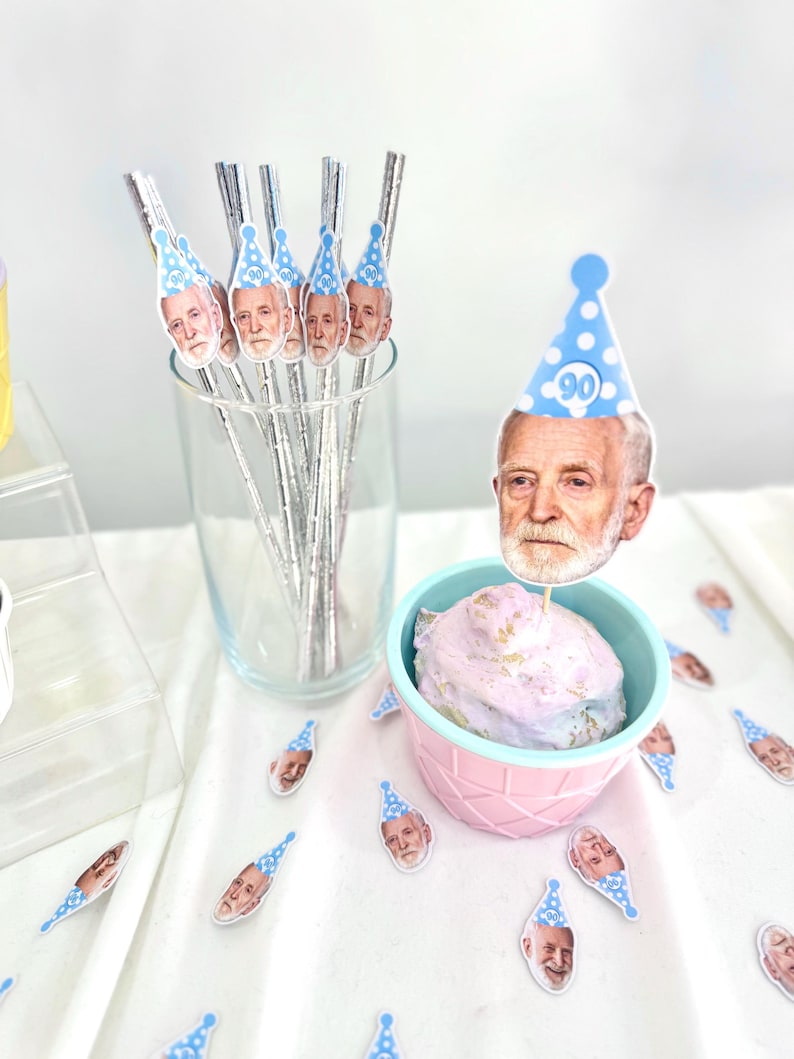 May include: Party decorations featuring a man's face wearing a blue party hat with the number 90. The decorations include drink stirrers in a glass, a cupcake with a topper, and scattered confetti.