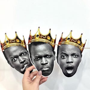 May include: A garland of three black and white face cutouts, each adorned with a gold crown. The faces display different expressions: one with a tongue out, one with a stern look, and one with an open mouth. The garland is strung on a thin twine.