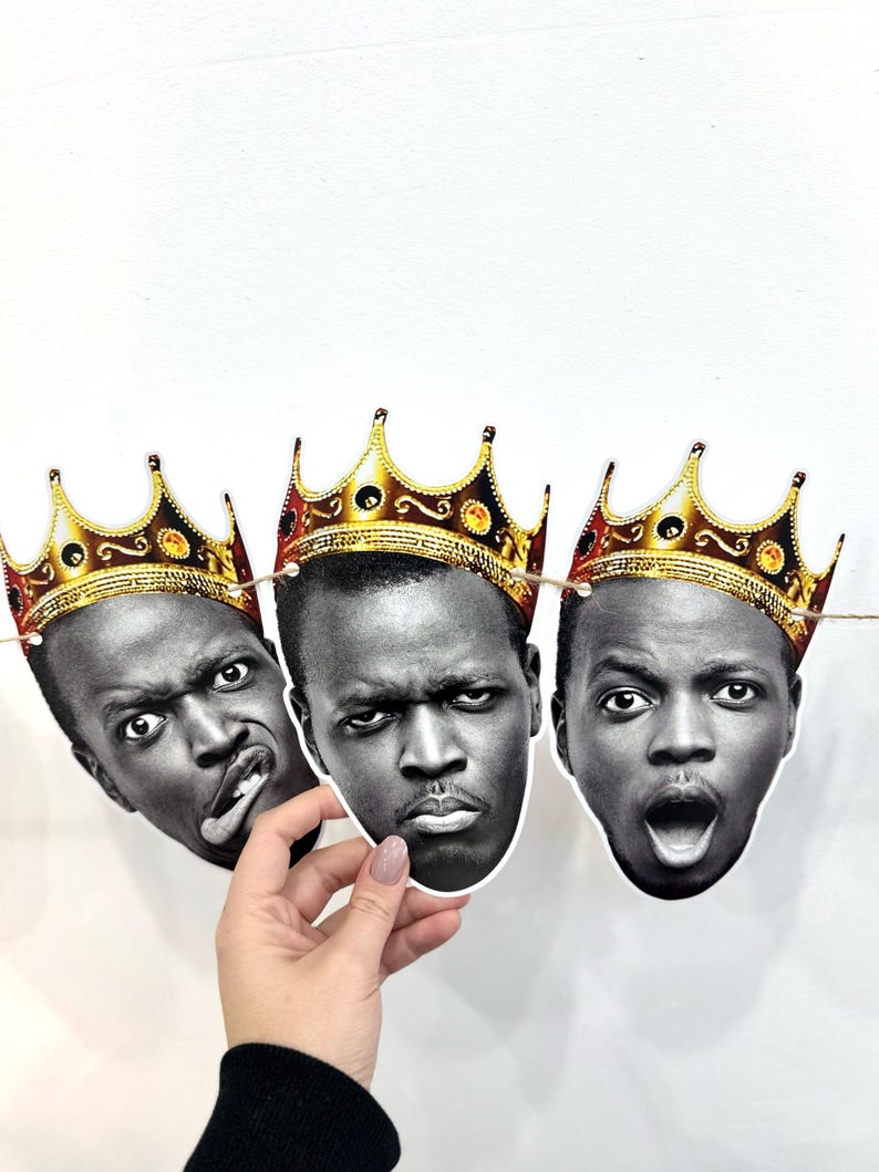 May include: A garland of three black and white face cutouts, each adorned with a gold crown. The faces display different expressions: one with a tongue out, one with a stern look, and one with an open mouth. The garland is strung on a thin twine.