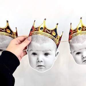 May include: A garland of baby face cutouts, each adorned with a gold-colored crown. The baby faces are in black and white, with a neutral background. The crowns have a detailed design with red and gold accents.