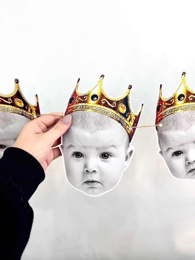May include: A garland of baby face cutouts, each adorned with a gold-colored crown. The baby faces are in black and white, with a neutral background. The crowns have a detailed design with red and gold accents.
