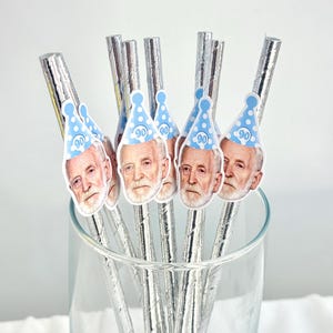 May include: A collection of silver-wrapped straws, each adorned with a sticker featuring a man's face and a blue party hat with the number 90. The straws are arranged inside a clear glass, suggesting a celebratory theme.