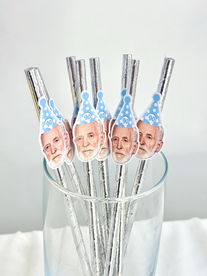 May include: A collection of silver-wrapped straws, each adorned with a sticker featuring a man's face and a blue party hat with the number 90. The straws are arranged inside a clear glass, suggesting a celebratory theme.
