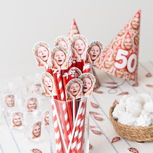 May include: A festive birthday party setup featuring straws, shot glasses, and a party hat, all adorned with a woman's face. Red and white striped straws are in a glass. A woven basket holds white meringues. A cake and a "50" hat are also visible.