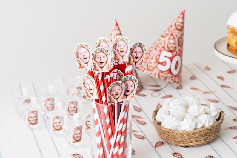 May include: A festive birthday party setup featuring straws, shot glasses, and a party hat, all adorned with a woman's face. Red and white striped straws are in a glass. A woven basket holds white meringues. A cake and a "50" hat are also visible.