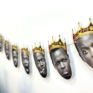 May include: A string of paper cutouts of a person's face wearing a gold crown. The faces have various expressions, including open mouths and sticking out tongues. The crowns are gold with red and black accents.