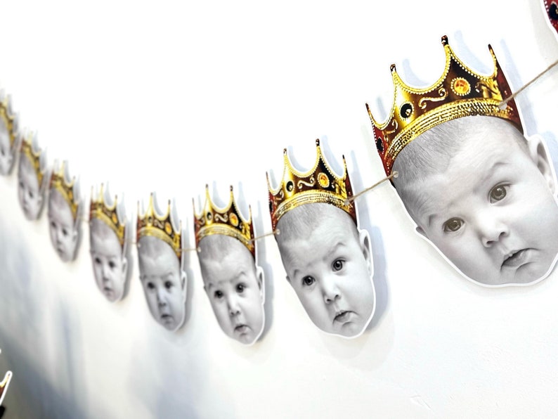 May include: A garland of baby faces with gold crowns. The black and white images of the babies are strung together with twine. The crowns are gold with red and black accents. The garland is hanging against a white wall.