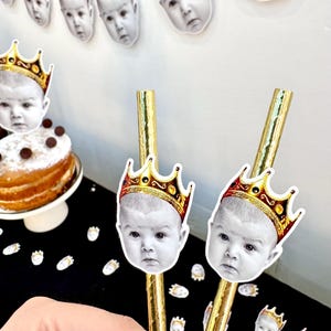 Op de afbeelding: Gouden rietjes met babygezichten met gouden kronen. Een banner met babygezichten met kronen hangt op de achtergrond. Een taart met chocoladeschilfers staat op een witte taartstandaard. Feestversieringen.
