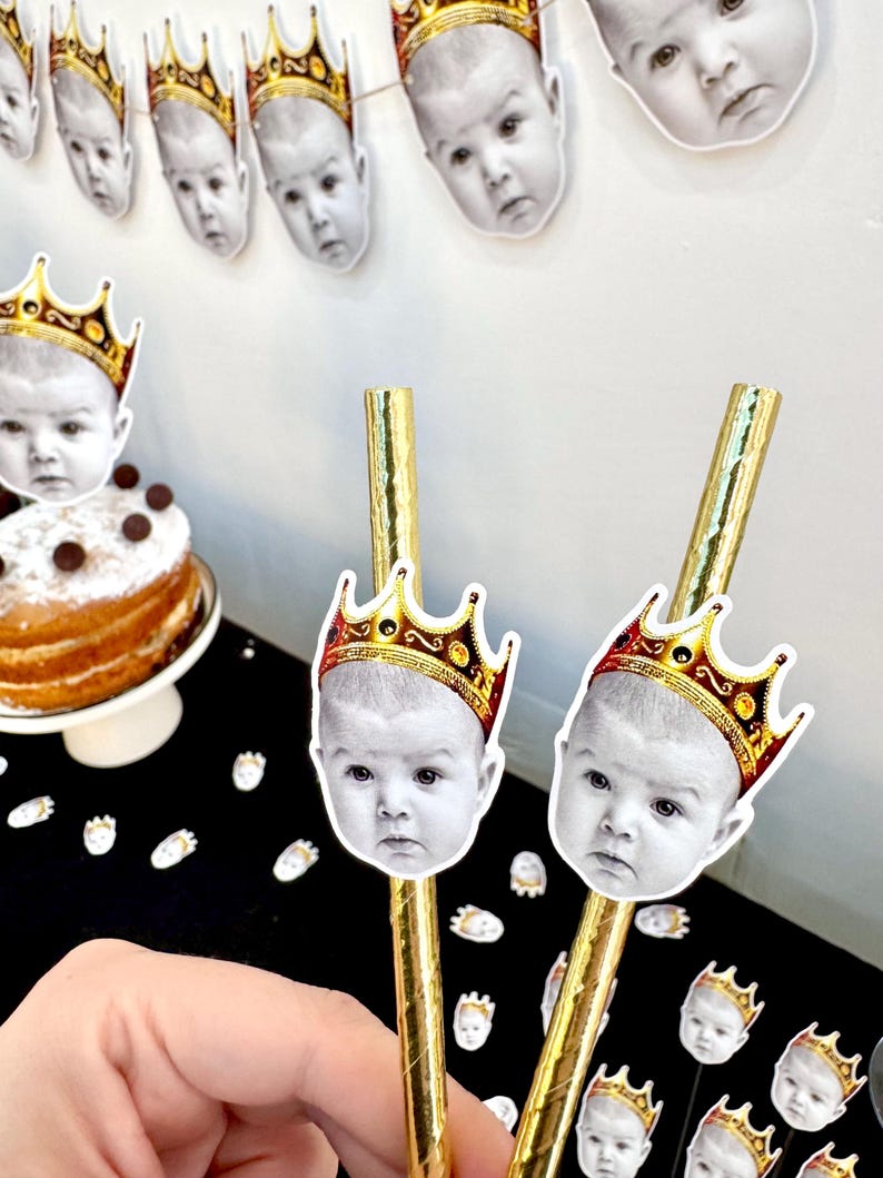 Op de afbeelding: Gouden rietjes met babygezichten met gouden kronen. Een banner met babygezichten met kronen hangt op de achtergrond. Een taart met chocoladeschilfers staat op een witte taartstandaard. Feestversieringen.