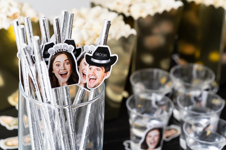 May include: A clear glass filled with silver straws topped with photo cutouts of people. Some cutouts say "Bride" and "Groom". Other items in the background include popcorn and clear plastic cups.