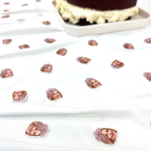 May include: A two-tier chocolate cake with white icing on a white plate. Small stickers of a woman's face wearing a tiara are scattered on a white surface. The stickers are pink and brown.