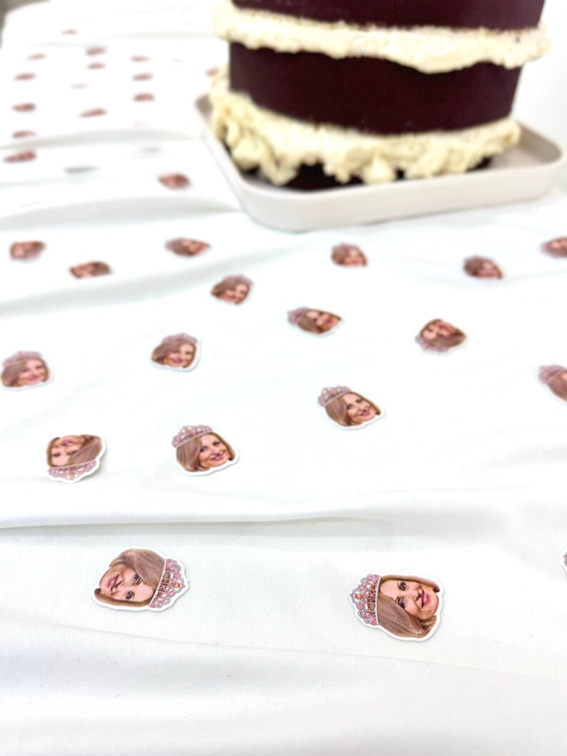 May include: A two-tier chocolate cake with white icing on a white plate. Small stickers of a woman's face wearing a tiara are scattered on a white surface. The stickers are pink and brown.