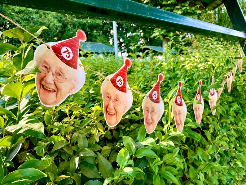 May include: A festive banner of smiling faces, each adorned with a red party hat featuring the number 75. The garland is strung against a backdrop of lush green foliage, perfect for a milestone birthday celebration.