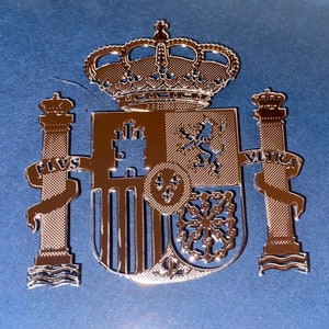 Puede incluir: Un emblema de metal dorado que representa el escudo de armas de España. El emblema incluye una corona, un escudo con un león, un castillo y una cadena, y dos pilares con las palabras "Plus Ultra" y "Ultra".