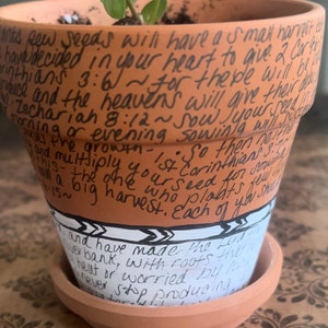 May include: A terracotta flower pot with a small plant growing inside. The pot is decorated with handwritten text, including bible verses and inspirational quotes. The text reads: "ants sew seeds will have a small harvest, have decided in your heart to give 2 Cor. 4:6, inthians 3:6~ for the be will be, auce and the heavens will give their d, Zechariah 8:12~Sow Jyoutu, morning or evening sowing will out, and multiply your seed for souring, this- the one who plaats, me growth. So then neithe, St Corinthians 3.7~, win a big harvest. Each of yo, -15~ Land have made the t, ver bank, with roots tharn, heat or worried by lond, never stop producing"