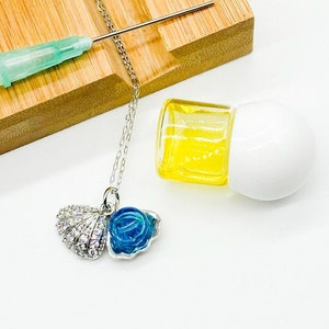 Puede incluir: Un collar de plata con un colgante de concha azul y blanca. El collar está sobre una superficie de madera con una botella transparente de líquido y una jeringa.