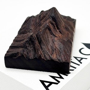 May include: A dark brown, rectangular wooden sculpture of a mountain range. The carving has detailed ridges and peaks, with a smooth, dark base. The sculpture is set against a white background.