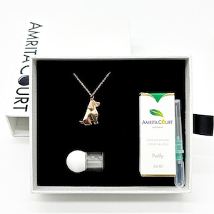 Peut inclure: Une boîte cadeau blanche avec un intérieur noir. La boîte contient un collier en or avec un pendentif en forme de chien origami, une petite bouteille d'huile essentielle étiquetée "Amrita Court Purify 10ml Net", et un compte-gouttes en verre.