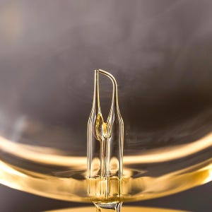 Op de afbeelding: Close-up van een helder glazen object met een uniek, langwerpig ontwerp. Het object heeft een gouden tint en staat tegen een vage, donkere achtergrond. Het ontwerp heeft een centraal, verticaal element met twee gebogen, symmetrische zijden.