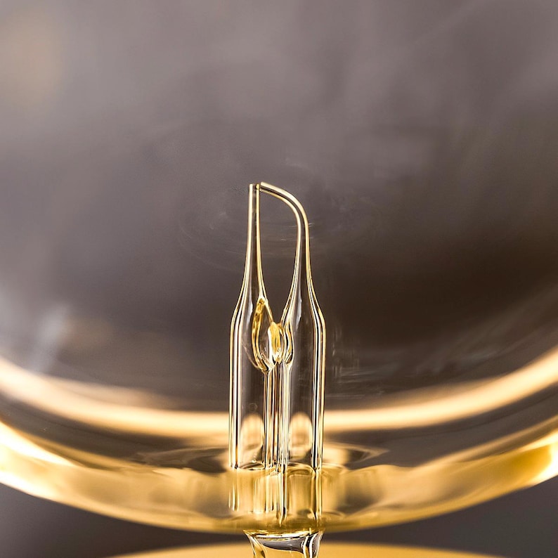 Op de afbeelding: Close-up van een helder glazen object met een uniek, langwerpig ontwerp. Het object heeft een gouden tint en staat tegen een vage, donkere achtergrond. Het ontwerp heeft een centraal, verticaal element met twee gebogen, symmetrische zijden.