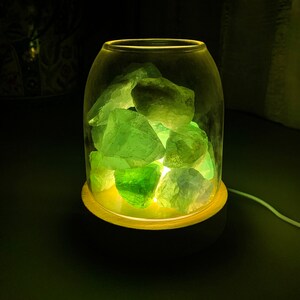 K&ouml;nnte beinhalten: Ein Glasgef&auml;&szlig; mit Holzsockel, gef&uuml;llt mit gr&uuml;nen Fluoritkristallen. Das Gef&auml;&szlig; wird von innen beleuchtet und l&auml;sst die Kristalle in warmem Licht erstrahlen.