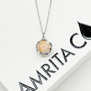 May include: A silver chain necklace with a delicate pendant featuring a gold and white orb encased in a silver cage-like design. The necklace is displayed on a white box with the word "AMRITA" printed in black.
