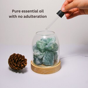 K&ouml;nnte beinhalten: Ein Glasgef&auml;&szlig; mit Holzsockel, gef&uuml;llt mit gr&uuml;nen Fluoritkristallen. Eine Hand h&auml;lt eine Tropfflasche und gibt &auml;therisches &Ouml;l in das Gef&auml;&szlig;. Der Text "Pure essential oil with no adulteration" ist &uuml;ber dem Gef&auml;&szlig; sichtbar.
