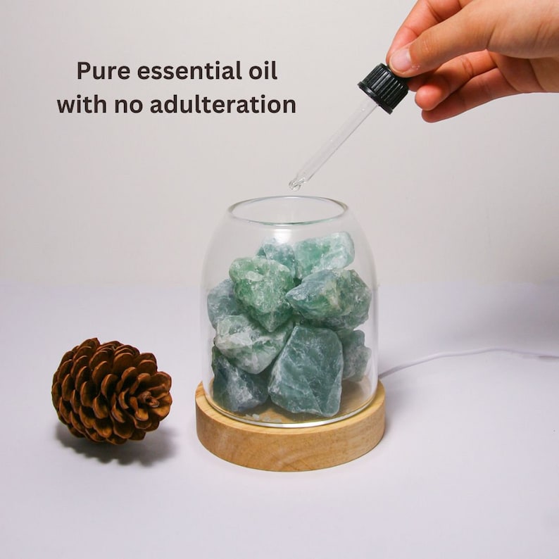 K&ouml;nnte beinhalten: Ein Glasgef&auml;&szlig; mit Holzsockel, gef&uuml;llt mit gr&uuml;nen Fluoritkristallen. Eine Hand h&auml;lt eine Tropfflasche und gibt &auml;therisches &Ouml;l in das Gef&auml;&szlig;. Der Text "Pure essential oil with no adulteration" ist &uuml;ber dem Gef&auml;&szlig; sichtbar.