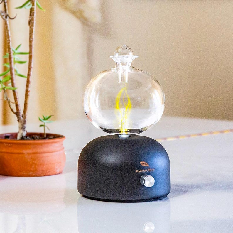 Op de afbeelding: Een zwarte etherische olie diffuser met een doorzichtige glazen bolvormige bovenkant. De bol heeft een vlamachtig geel licht aan de binnenkant. De basis heeft een zilveren knop en de tekst "Ambiance". Een kleine plant in een pot staat op de achtergrond.