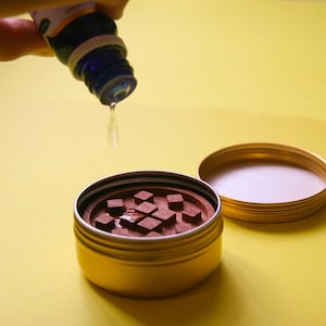 May include: A gold metal container with a wooden insert filled with small squares. Essential oil is being poured into the container.