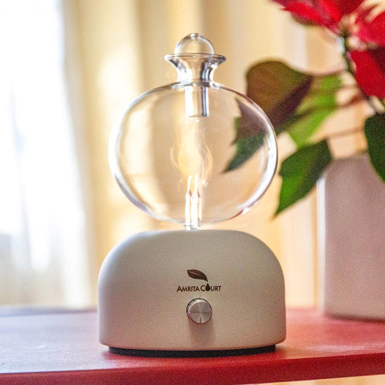 Op de afbeelding: Een heldere glazen etherische olie diffuser met een bolvormige bovenkant en een lichtgrijze basis. De basis heeft een zilveren knop en de tekst "AMRITA COURT". De diffuser staat op een rode ondergrond.
