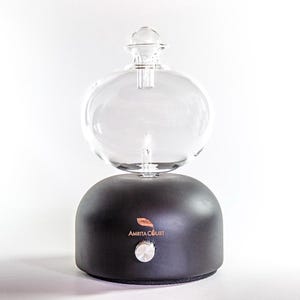 Op de afbeelding: Een heldere glazen etherische olie diffuser met een afgeronde bovenkant en een zwarte basis. De basis heeft een zilveren knop en de woorden "Amrita Court" erop gedrukt. De diffuser is ontworpen om etherische oli&euml;n te verspreiden.