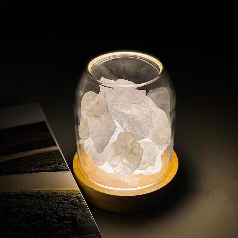 K&ouml;nnte beinhalten: Eine wei&szlig;e Kristallsteine-Lampe mit einem klaren Glasgef&auml;&szlig; und einem Holzsockel. Die Lampe ist eingeschaltet und die Kristalle leuchten.