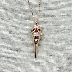 Peut inclure: Un collier en or rose avec un pendentif orné et détaillé en forme de larme pointue et allongée. Le pendentif est décoré de petites pierres claires.