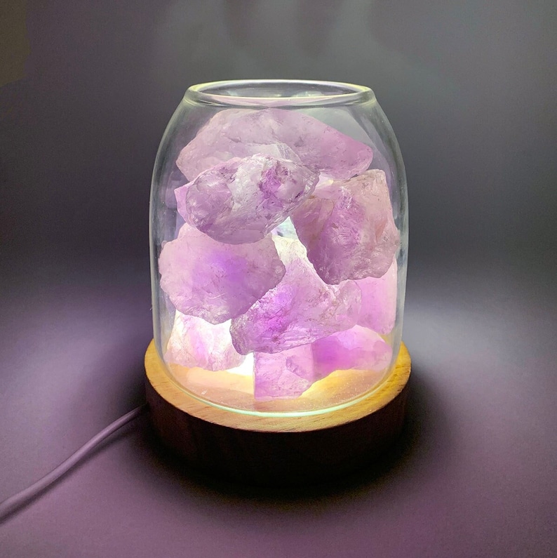 K&ouml;nnte beinhalten: Eine Glaslampenbasis mit einem Holzboden, gef&uuml;llt mit Amethystkristallen. Die Lampe ist eingeschaltet und strahlt ein sanftes violettes Licht aus.