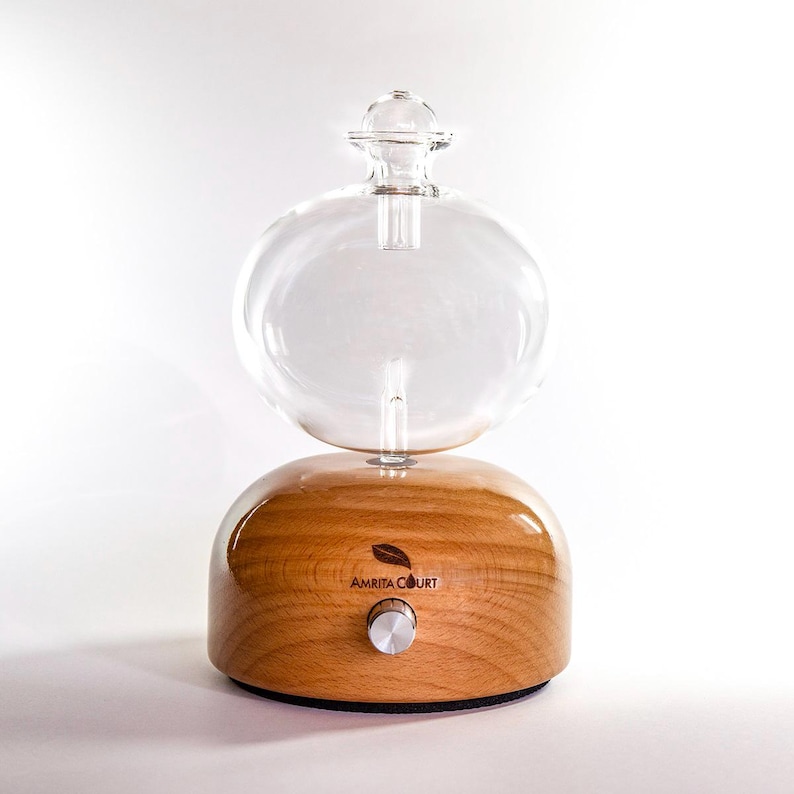Op de afbeelding: Een heldere glazen etherische olie diffuser met een afgeronde bovenkant en een houten basis. De basis heeft een zilveren knop en de tekst "AMRITA COURT". De diffuser is ontworpen om etherische oli&euml;n te verspreiden.